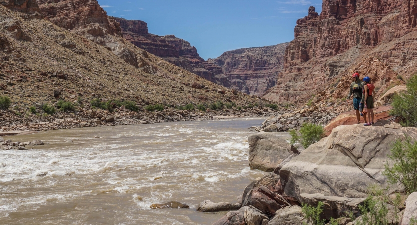 Two people stand on a rock overlooking a river below. There are tall canyon walls on either side of the river.